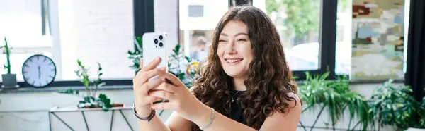 stock image A plus-size professional with long curly hair smiles while taking a selfie in a lively office space filled with greenery.