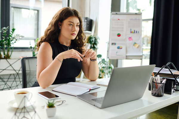 A confident plus-size woman with long curly hair focuses on her laptop in a bright office, surrounded by plants.