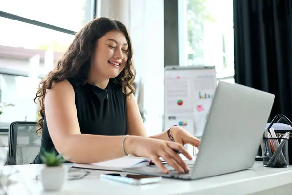 stock image A young, confident plus size woman with long curls engages in her work at a bright office, radiating positivity.