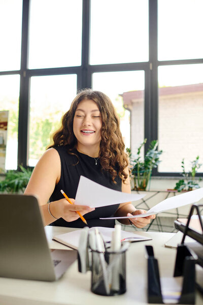 A radiant plus-size woman with curly hair engages in her work, sorting through documents with enthusiasm in a sunlit office.