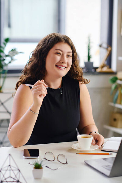 Уверенная в себе молодая женщина с вьющимися волосами занимается своей работой, наслаждаясь чашкой кофе за своим столом.