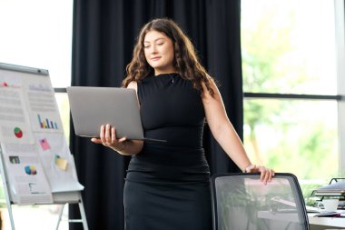 Kendine güvenen, uzun kıvırcık saçlı iri yarı bir kadın şık bir ofis ortamında laptopuyla aktif olarak iş birliği yapıyor..