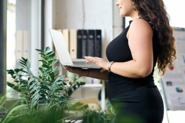 Profesyonel ve cüsseli bir kadın, bitkilerle zenginleştirilmiş canlı bir ofis atmosferinde dizüstü bilgisayarıyla güvenle ayakta duruyor..