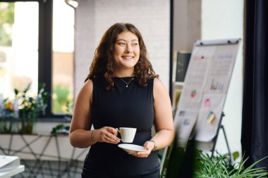 A young and beautiful plus size woman with long curly hair smiles while enjoying a coffee break at work. clipart