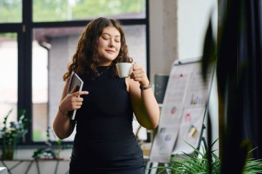 A young plus size woman with long, curly hair savors her coffee while engaged in office tasks at a stylish workplace. clipart