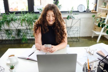Canlı bir ofis ortamında, kıvırcık saçlı iri yarı bir kadın masasında çalışırken telefonunu neşeyle kullanır..