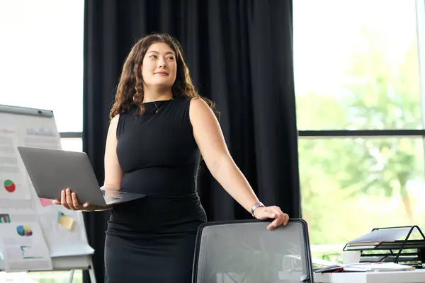 stock image A confident young woman with curly hair stands in a sleek office, engaging in business tasks while holding a laptop.