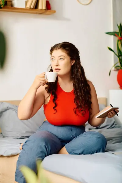 stock image A young plus-size woman with curly hair sits on her bed, sipping tea and jotting down thoughts in her journal, embracing a tranquil moment.