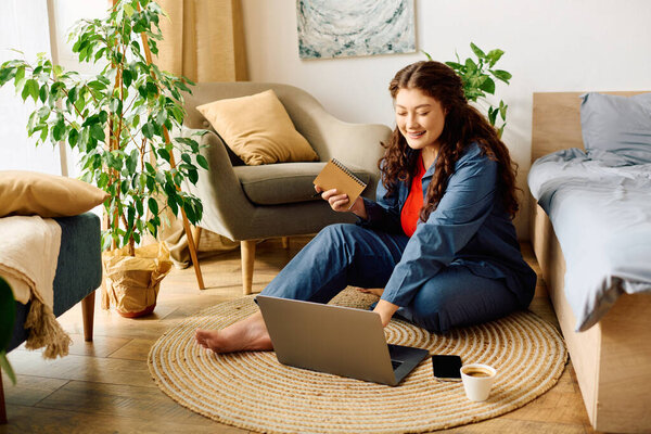 Веселая женщина plus size удобно сидит на полу, занимаясь ноутбуком и делая заметки с улыбкой.