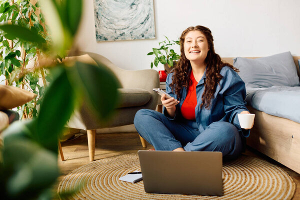 Веселая женщина plus size с кудрявыми волосами сидит на полу, держит чашку кофе и смотрит на свой телефон в уютном пространстве.