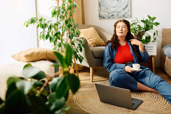 Молодая красивая женщина plus size сидит дома на полу, пьет чай и наслаждается своей уютной жизнью..
