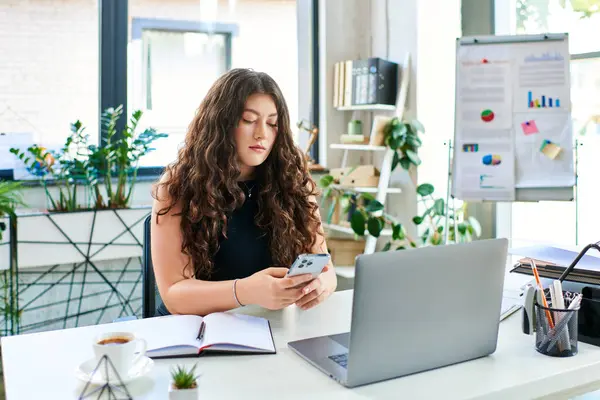 stock image A confident plus size woman with long curly hair multitasks in a modern office, focusing on her smartphone while working.