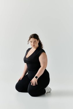 Güzel, iri yarı, siyah giyinmiş bir kadın zarif bir şekilde diz çöker, kendine güven ve zarafet saçar..