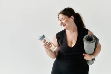 Kadın, elinde su şişesi ve yoga minderiyle pratik yapmaya hazır bir şekilde gülümsüyor..