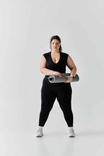 Уверенная женщина plus size стоит высокая с ковриком для йоги, готовая к практике в ярком пространстве.
