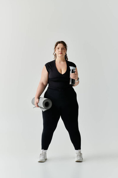 Изящный плюс размер женщина стоит уравновешенный с циновкой йоги и бутылкой воды, готовый принять фитнес.