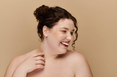 Kıvırcık saçlı, iri yarı bir kadın tarafsız bir arkaplanda neşe ve güven yayar..