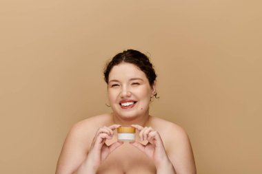 A confident and beautiful plus size woman smiles brightly while holding a skincare product. clipart