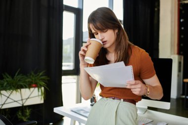 Modern bir ofiste, genç bir ikili olmayan kişi, elinde kahveyle kağıtları incelikle inceler..