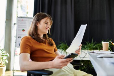 İkili olmayan bir kişi, bitkilerle dolu bir ofis alanında dinlenirken belgeleri gözden geçirir.