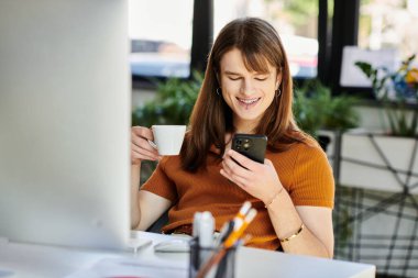 A cheerful non binary person relaxes with coffee, engaged with their smartphone in a bright office.