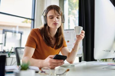 Çağdaş bir ofiste, ikili olmayan bir birey kahvesini yudumlar ve akıllı telefonlarını kontrol eder..