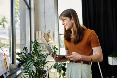 A young non binary person smiles while working on a laptop, surrounded by plants in a bright office. clipart