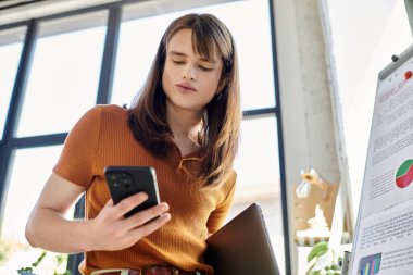 Ofis dekoru ve bitkilerle çevrili genç, ikili olmayan bir kişi telefonunu inceliyor..