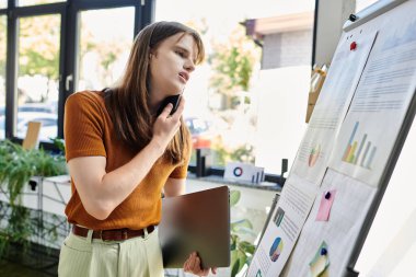 In a vibrant workspace, a non binary person reflects on charts while holding a tablet. clipart