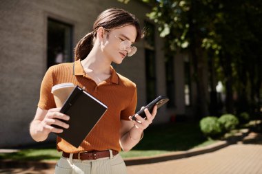 Genç, ikili olmayan kişi dışarıda duruyor, telefonuna odaklanmış, elinde kahveyle..