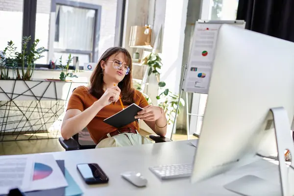 stock image A young non binary individual thoughtfully reflects while surrounded by greenery and office tools.