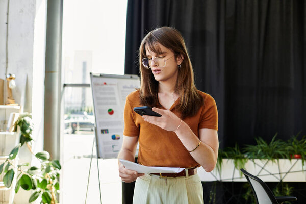 Молодой не двоичный человек рассматривает заметки при использовании смартфона в ярком офисе.