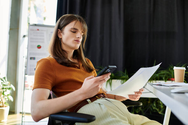 A young non binary person relaxes in an office, reviewing papers and using their phone.
