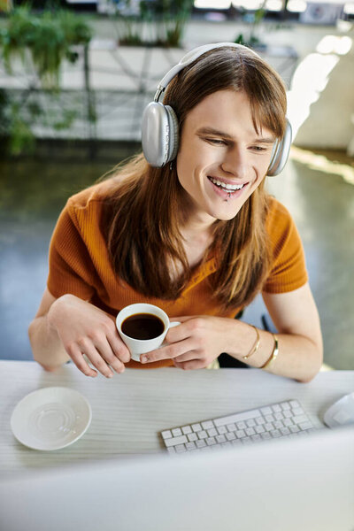 Молодой небинарный человек улыбается, потягивая кофе в ярком офисе.