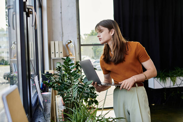 A young non binary individual holds a laptop while looking out the window in a vibrant workspace.