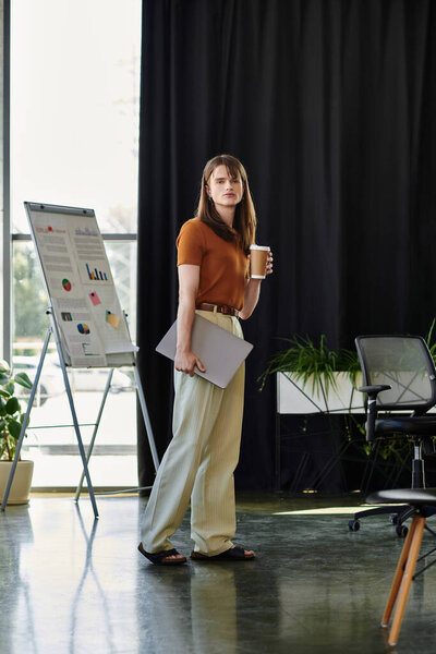 In the office, a non binary individual holds a coffee cup while looking thoughtful and engaged.