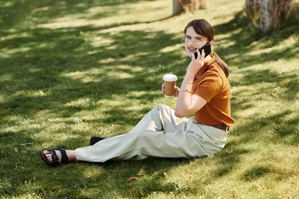 stock image A young non binary person sits on the grass, chatting lively on the phone.