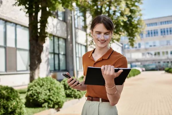 stock image A young non binary person smiles while reviewing notes outdoors, surrounded by greenery.