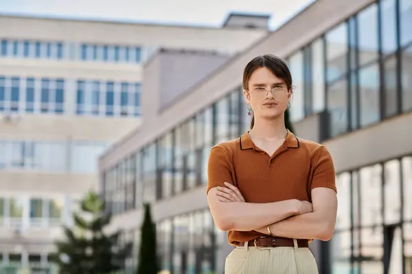 stock image A young non binary person stands confidently with crossed arms in a bright office area.