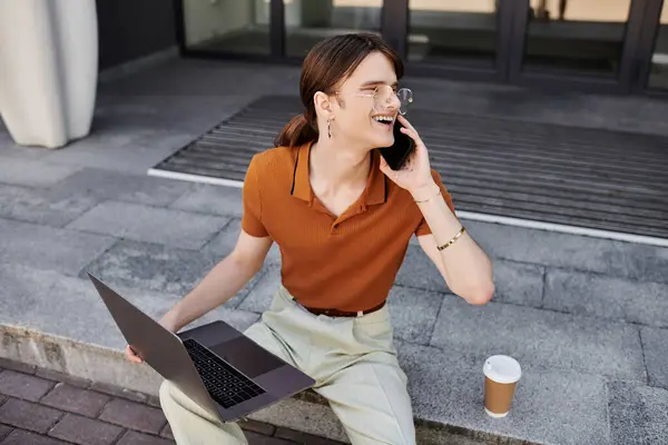 stock image A young non binary individual laughs while talking on the phone, balancing work and play outdoors.