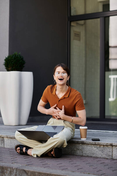 A young non binary person happily works on their laptop while sipping coffee on the steps.