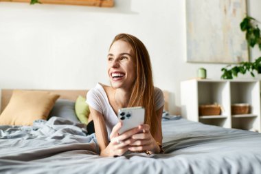 Genç bir kadın yatağında dinlenirken gülümsüyor, telefonunu tutuyor ve bir anlık mutluluğu kucaklıyor..