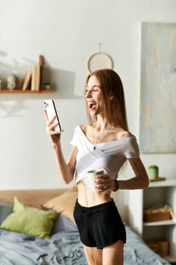 Neşeli bir suratı olan genç bir kadın, telefonunu kullanırken bardaktan yudum yudumlayarak, evde gününün tadını çıkarıyor..