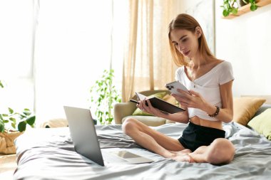 Genç bir kadın, mutlu bir şekilde yatağında oturmuş ev atmosferiyle çevrili telefon ve defterini dengeliyor..