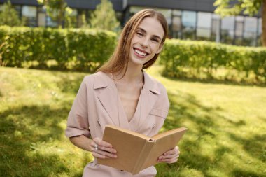 A young woman with anorexia smiles brightly as she spends a sunny afternoon outdoors, reading in a lush green setting.