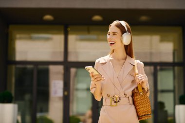 İştah açıcı bir gülüşü olan genç bir kadın açık havada, kulaklık takıyor ve telefonunu kontrol ederken mutlu oluyor..