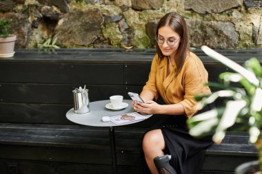 Protez bacaklı genç bir kadın rahat bir kafede rahatlar, akıllı telefonuyla meşgul olur ve kahvesini yudumlar..