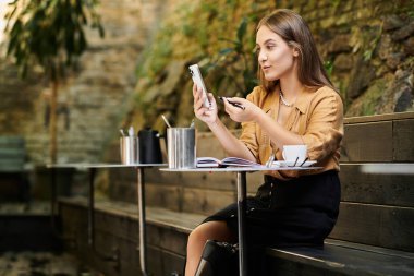 In a cozy cafe, a young woman confidently navigates her day, interacting with her phone while seated at a table. clipart