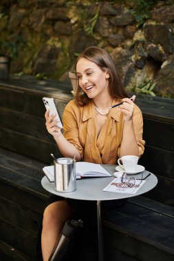 Protez bacaklı genç bir kadın bir kafede dinleniyor, telefonuna gülümsüyor ve kahvesinin tadını çıkarıyor..