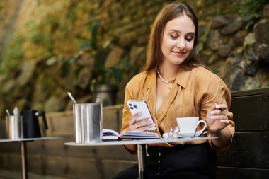 A young woman with a prosthetic leg smiles while sipping tea in a cozy cafe, immersed in her phone and surroundings. clipart